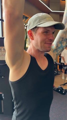 A man in a black tank top and a light grey corduroy baseball cap is working out at the gym. He has blonde hair, blue eyes, and tattoos on his arm. He is smiling while lifting weights using a cable machine. Other gym equipment is visible in the background, as well as another person. The ceiling has reflective surfaces with warm lighting.