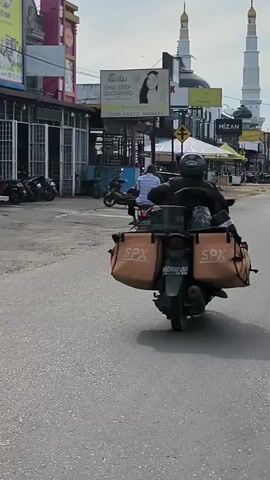 The video shows a rear view of a person riding a motorcycle on a paved road. The motorcycle has two large orange bags attached to the back, each with the SPX logo. The rider is wearing a helmet and a dark jacket. In the background, there are buildings, billboards, and a traffic light. Two domed structures, possibly minarets of a mosque, are visible in the distance. Another motorcycle is seen ahead of the rider. The environment appears to be a commercial area with shops and stalls lining the street. The road surface is asphalt and shows signs of wear. The lighting suggests it is daytime.