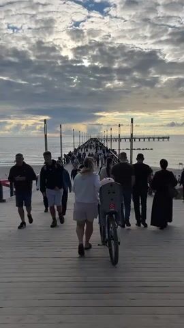 A low-angle shot shows a crowded wooden pier extending into the ocean under a cloudy sky. People are walking in both directions on the pier, some pushing strollers and one person riding a bicycle with a child seat. The sun is partially visible through the clouds, casting light on the water. The overall mood is one of leisure and activity.