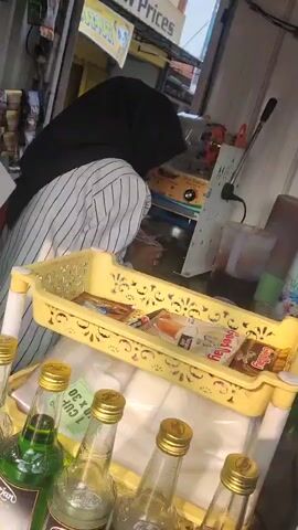 The video shows a person wearing a black hijab and a striped shirt in what appears to be a small street-side stall. They are leaning over a counter, seemingly preparing something. In the foreground, there is a yellow plastic basket containing various packaged snacks. Below the basket, several glass bottles with gold caps are visible, some containing green liquid. In the background, there are shelves with more products, a sealing machine, and a large jug. Outside the stall, a street scene is visible with people, motorcycles, and a vehicle. A sign with "Low Prices" is also visible.
