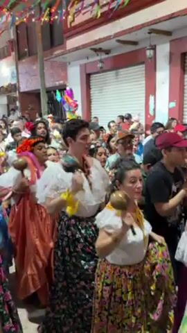 A lively street parade is in progress, filled with people celebrating under colorful overhead decorations. The participants are dressed in a mix of casual and traditional clothing, with several women wearing floral skirts, white blouses, and flower headpieces. They're holding maracas and other objects, shaking and dancing along to the music, though the source isn't visible. The crowd includes a diverse group of people, many smiling and appearing to be engaged in the festivities. Buildings line the street, some with closed shutters. The overall atmosphere is cheerful and celebratory.