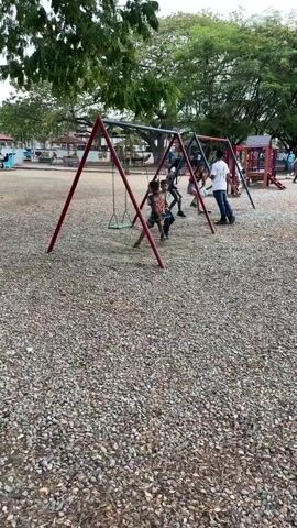 The scene is a playground, covered with small rocks, where several people are on swings. There are three swingsets visible, each with red A-frame supports and a dark crossbar. People of varying ages are on the swings, including children and adults.  One person is standing near a swing, seemingly assisting. In the background, there's a structure resembling a gazebo or pavilion, along with other playground equipment. Trees surround the area, casting shadows on the ground. Other people are visible in the background, moving about the park.  The overall atmosphere appears relaxed and casual.