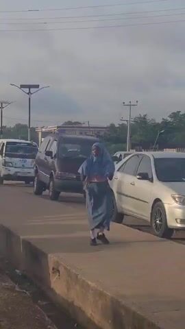 A young person wearing a light blue hijab and long blue skirt is walking alongside a road with traffic. The road is lined with cars and a van.  Behind the cars is a business sign. A tall, metal light post with solar panels is visible. Palm trees and power lines are also visible. The sky is overcast.