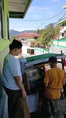 A young male wearing a white t-shirt, brown sarong, and black peci looks to his left and then turns to face the camera. Next to him, another young male wearing a yellow t-shirt and patterned sarong stands with his back to the camera, facing a food cart. The food cart has a glass display case filled with food items and some containers. A green tarp is draped over the top of the cart. The background shows a building with white and green trim, a stone wall, and trees and hills in the distance. The scene appears to be outdoors during the day with bright sunlight.