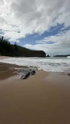 The video shows a beach with tan sand and waves coming ashore. A dark rock sits in the sand near the bottom left corner. In the distance, a green cliff covered in trees curves towards the ocean. The sky is partially cloudy with patches of blue. The overall lighting is bright.