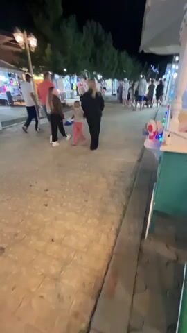 The video shows a nighttime scene on a cobblestone street lined with shops and market stalls. Several people are walking along the street, some in groups and others individually. The lighting is dim, with streetlights and lights from the shops illuminating the area. In the foreground, a person's hand is visible holding a black object. Further down the street, a family appears to be walking together, with two adults and two children. One of the adults is carrying a small child. Another person in a light blue shirt and orange shorts walks past a sign with images of food. The overall atmosphere seems relaxed and casual, with people enjoying an evening stroll and browsing the shops.