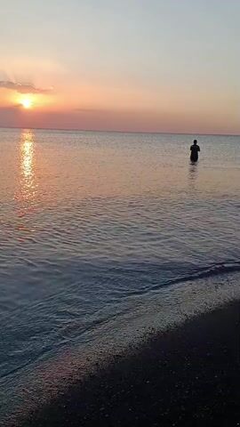 The video shows a serene ocean scene at sunset. The sun is low on the horizon, casting a warm, orange glow across the water and sky. Gentle waves lap at the shore, which is dark and pebbly. In the distance, a lone figure stands in the shallow water, silhouetted against the setting sun. The overall mood is peaceful and contemplative.