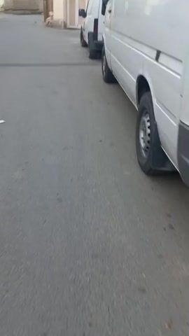 This video appears to capture a brief, unedited street-level view of a residential or light industrial area in Khujand, Tajikistan. The scene is dominated by several white vans parked alongside a paved road. The perspective is low, close to the ground, and the camera is moving, suggesting it might be attached to a vehicle or being held by someone walking.

The vans are the primary objects, with one white van prominently featured in the foreground, its side facing the camera. Another white van is visible behind it, and further down the street, a third van is partially obscured. The vans appear functional, with one having a roof rack.

The setting is a street with buildings on either side. The architecture of the buildings is varied, with some appearing to be older, weathered structures with visible signs of wear and tear, including boarded-up windows and exposed brickwork. One building has a black tarp covering a section of its facade. The road itself is asphalt and shows some wear.

The time of day appears to be overcast, possibly late afternoon or early morning, given the diffused light. There are no clear indicators of specific weather conditions beyond the overcast sky. The overall mood is ordinary, depicting a mundane street scene. No people are clearly visible in the frame. There is no visible text within the media file. The activity taking place is simply the movement along a street where vehicles are parked.
