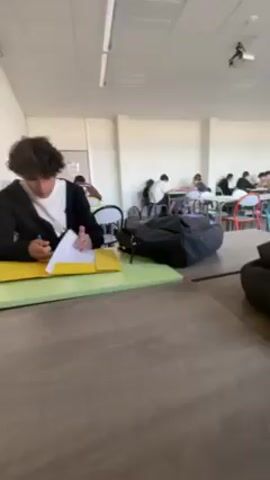 Here is a concise description of the video:

The video shows a classroom setting. Several students are sitting at desks working, most likely taking a test or doing individual work. Some are visible, while others are partially obscured by bags on the tables. The classroom has fluorescent lighting and a projector hanging from the ceiling. The general atmosphere seems focused and quiet.