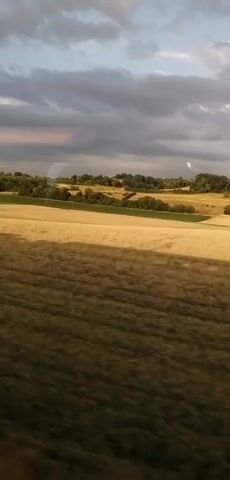 The video shows a view from a moving vehicle, likely a train, looking out over a rural landscape. The primary subjects are the fields, rolling hills, and trees that make up the setting. The fields appear to be golden, suggesting a harvest season, and are interspersed with patches of darker vegetation. The sky is filled with dramatic, grey clouds, hinting at an overcast or approaching storm, though there are also breaks of sunlight casting shadows across the fields. The time of day appears to be late afternoon or early evening, given the quality of the light. The location is identified as Bavois, Switzerland. The overall mood is serene and expansive, capturing the beauty of the countryside as it passes by. There are no visible people, objects, or text in the video. The activity is simply the passage through the landscape.