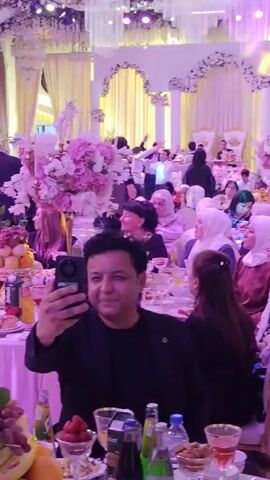This video shows a wedding reception or similar formal gathering inside a large, ornate hall. Guests are seated at round tables laden with food and drinks. The hall is decorated with white drapes, floral arrangements, and elaborate chandeliers. In the foreground, a man in a black shirt is taking a selfie with his phone. In the background, a group of musicians in matching blue and black outfits are playing large, horn-like instruments. Several people are standing and dancing in the background. The lighting is a mix of white and purple, creating a festive atmosphere.