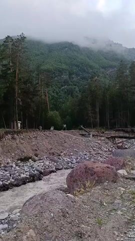 A drone shot begins in a rocky, debris-strewn riverbed with murky, fast-flowing water. Trees line the banks, with a dense forest stretching up a steep, fog-shrouded mountain in the background. Several large rocks are scattered in the foreground, with smaller rocks and dirt making up the riverbed. A narrow wooden bridge crosses the river further upstream, partially obscured by trees.