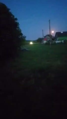 The video shows a rural landscape at dusk or night. In the foreground, there is a dark, out-of-focus area, likely foliage. In the mid-ground, there is a grassy field with a few scattered objects. Further back, there are several buildings, possibly houses, with some lights on. A tall pole with what appears to be a light fixture on top stands between the buildings. There is a bright, circular object in the sky, possibly the moon or a strong light source. In the distance, a vehicle's headlights are visible. The sky is a deep blue, indicating low light conditions. The overall impression is a quiet, rural setting at twilight or after dark.