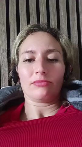 A woman with blonde hair and headphones is in the center of the frame, wearing a red shirt. She is lying down and appears to be recording a video of herself. She makes a few facial expressions. The background is a wall with vertical wooden slats.