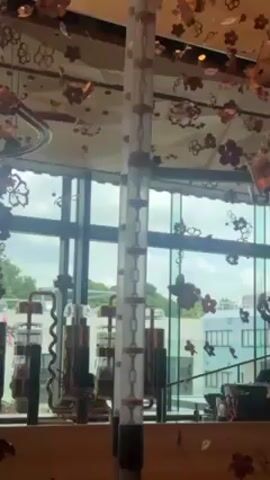 The video presents an interior shot, likely within a café or similar establishment, showcasing distinctive design elements. A tall, transparent cylindrical structure with chain-like decorations stands prominently in the foreground. The space is adorned with numerous metallic flower-shaped ornaments hanging from the ceiling, creating a floral-themed ambiance. A large glass window offers a view of an urban landscape featuring buildings and lush greenery, with an overcast sky.