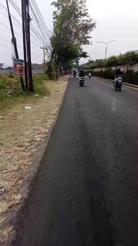 A video taken from a moving vehicle shows a paved road with motorcycles and trees on the side. The road is dark and appears wet. There are several motorcycles on the road, some with riders. Trees line the left side of the road, and there are buildings in the background. The sky is overcast. The video appears to be taken during the day.