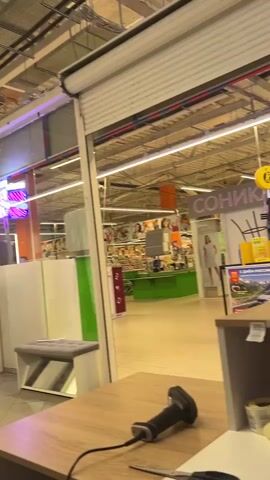 This media file appears to be a video taken inside a retail store, possibly a supermarket or department store. The camera is positioned at a checkout counter, with a barcode scanner visible in the foreground. Through an open doorway, the interior of the store is visible.

**Subjects:**
*   **People:** Several people are visible in the store, walking and shopping. One person, a woman in a light-colored dress, is standing near a display with the word "СОНИКА" visible. Another man and woman are walking down an aisle. A cardboard cutout of a man in a suit is also present.
*   **Objects:** A barcode scanner is in the foreground. In the store, there are shopping baskets, shelves stocked with merchandise, displays, and signage.
*   **Scene/Setting:** The setting is an indoor retail environment, characterized by bright fluorescent lighting, high ceilings with exposed infrastructure, and rows of merchandise. The overall impression is of a busy commercial space.

**Activity or Event:**
The primary activity is shopping. People are browsing or moving through the store. The presence of the barcode scanner suggests that a transaction is either about to occur or has just concluded.

**Notable Details:**
*   **Time of Day/Weather:** No direct indicators of time of day or weather are present, as it is an indoor scene.
*   **Location Cues:** The visible Russian text on signage suggests the location is in Russia. Specifically, one sign reads "СОНИКА", another has "FLOWERS" and "OZON" (a Russian e-commerce company). The context provided states the location is Cherepovets, Russia.
*   **Emotions:** No discernible emotions are evident from the brief glimpses of people. The atmosphere appears to be typical of a retail environment.

**Visible Text:**
*   "СОНИКА"
*   "FLOWERS"
*   "OZON"
*   "С ДНЁМ РОССИИ!" (Happy Russia Day!) - This is on a poster depicting a landscape.
*   Additional text in Cyrillic script is visible on various signs, but is not clearly legible.