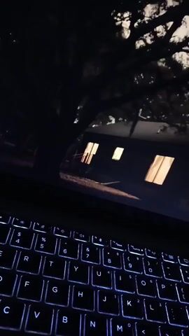 The video displays a close-up view of a laptop keyboard with illuminated keys. The screen of the laptop shows an image of a dark, nighttime scene with several windows emitting a warm, orange light, suggesting interior illumination. Trees and their branches are visible against a faintly lit sky, casting shadows over the scene. The image on the screen appears to be a still photograph or a paused video frame. The overall ambiance is dark and somewhat eerie, possibly depicting a residential building or a structure at night. There are no people or active movements visible in the video.