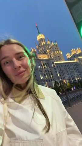 A young woman with light brown hair and blue eyes is taking a selfie-style video. She is wearing a white jacket and has some bracelets on her wrist. In the background, there is a large, brightly lit building with multiple spires and a green traffic light. The sky is a deep blue, suggesting it is either dusk or dawn. The camera is tilted at an angle, and the woman is looking directly into the lens and smiling slightly. She is running her hand through her hair. The overall scene appears to be taken in an urban environment.