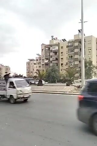 The video shows a street scene in what appears to be an urban area. Several multistory apartment buildings line the background under a cloudy sky. The street is active with vehicles. Initially, a small truck with people on top and a motorcyclist pass by. Following this, a convoy of cars drives through, some decorated with green and white flags. A man stands on the side of the road, observing the passing vehicles with his arms crossed. The general impression is a celebratory or festive atmosphere with the vehicular procession.