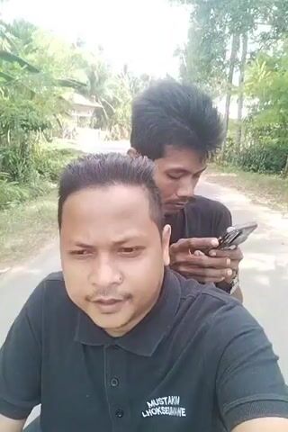 Two men are riding on a motorcycle outdoors. The man in the foreground, who is closer to the camera, is wearing a black polo shirt. He has dark hair styled with a side part and is looking directly at the camera with a serious expression. The man behind him is wearing a black t-shirt and has spiky dark hair. He is looking down at a smartphone he is holding in his hands. The motorcycle is on a dirt road that is lined with trees and vegetation. The overall environment appears to be a rural or tropical setting, with lush greenery visible in the background and a bright sky. The video is a selfie-style recording, likely taken by the man in front.