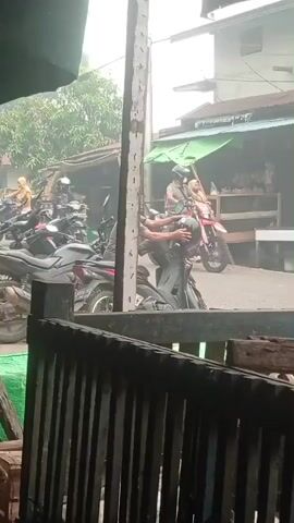 A person wearing a grey t-shirt and shorts is standing on the left side of the frame next to a parked motorcycle. Another person, wearing a t-shirt and helmet, is sitting on a black motorcycle in the center of the frame. A red motorcycle is visible on the right side of the frame, with a person wearing a helmet and mask riding it. Several other motorcycles are parked in the background. The environment appears to be a street with buildings and trees. The lighting is bright, suggesting it is daytime.