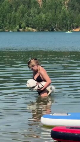 A woman in a black bikini is standing in a lake holding a small, white dog in her arms. The water reaches about her waist. The dog is wearing a harness. The woman is also wearing sunglasses.  Behind them is a lake surrounded by trees. In the foreground are two colorful inflatable objects. The weather appears sunny.