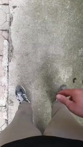 A person wearing grey sneakers and tan pants is standing on a concrete path next to a tiled walkway. The camera is positioned from the person's perspective, looking down at their legs and a black dog with brown markings. The dog is a Rottweiler and is lying on its back at the person's feet. The dog is panting and has its mouth open, with its tongue visible. It appears to be happy and playful, looking up at the camera with its eyes. The scene is outdoors, and the ground is a rough, grey concrete. There are some small brown objects on the ground to the right of the dog, which appear to be pebbles or dried leaves. The lighting suggests it is daytime. The video is a short clip, showing the dog's playful behavior.