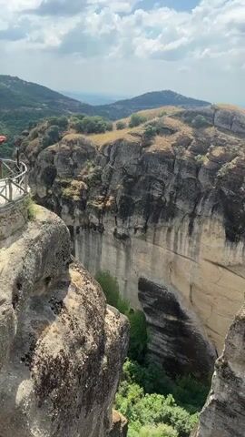 A vertical video pans from left to right, revealing a dramatic landscape dominated by towering rock formations. The foreground consists of rough, textured rock surfaces, some covered in moss. The middle ground showcases immense, sheer cliffs with vertical striations, carved by time and elements. Lush green vegetation, including trees and bushes, grows in the crevices and at the base of the cliffs. In the distance, a village with red-tiled roofs is nestled among rolling hills and fields. Mountains form the backdrop, with a hazy sky dotted with white clouds. The overall impression is one of natural grandeur and isolation.