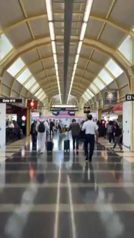 The video shows the interior of an airport terminal or walkway. Several people are walking in both directions along a polished floor. Many are pulling luggage behind them. The structure overhead is arched with rows of rectangular lights. On either side of the walkway are storefronts or areas with seating. Signs indicating gate information are visible above the people.