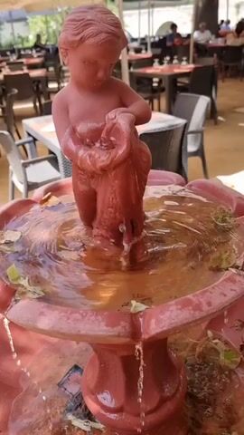 A video shows a red fountain statue of a cherub holding a bird. Water flows from the statue and over the edge of the fountain. In the background, patrons sit at tables under umbrellas at an outdoor restaurant.