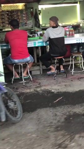 The video shows two men seated on stools at a table in what appears to be an outdoor food stall or small restaurant. The man on the left is wearing a red shirt and blue shorts, facing away from the camera. The man on the right is wearing a grey and black hooded sweatshirt and black pants, also facing away from the camera. Both men are looking towards the table, suggesting they are eating or ordering food. There are various items on the table and shelves behind them, including bottles, food items, and what looks like merchandise. A motorcycle is partially visible in the foreground on the left side of the frame, indicating a street-level or roadside setting. The ground is paved with a mix of asphalt and possibly brick or tile.