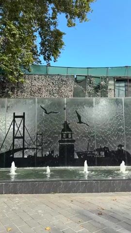 The video begins with a wide shot of a fountain with a water wall featuring a black silhouette of an Istanbul skyline including the Bosphorus Bridge, the Maiden's Tower, and ships. Small jets of water spray from the fountain in front of the water wall. Above the water wall, the top of a stone wall covered with camouflage netting is visible, with trees and a blue sky beyond. The camera pans to the right, revealing more of the Istanbul skyline silhouette on the water wall, including a mosque and people. A building with a modern facade, featuring balconies and advertising for "GHAFET MUSTAPHA INN," is visible behind the water wall, with a green vertical garden on its side. Several people are walking along a paved walkway in front of the fountain and building. Two men are walking towards the camera, each carrying shopping bags. The video ends with a view of the street scene and the building.