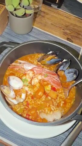 A close-up, overhead shot of a seafood paella dish served in a black, shallow pan with two handles. The paella is a vibrant orange-red, with visible grains of rice, chunks of vegetables like peppers, and assorted seafood. Prominently featured are two large, pink prawns arranged on top, their heads facing left. Several dark blue mussels, some open and some closed, are scattered around the prawns and within the rice. Two white clams with visible flesh are positioned on the left side of the dish. A spoon with a silver handle is partially submerged in the paella on the right side. The pan sits on a white plate, which is placed on a blue and grey striped placemat. In the background, a small potted succulent plant with green leaves is visible in a grey pot. To the left of the plant is a brown wooden object with carved patterns and a QR code. Further back, a wooden table surface is visible, along with a salt and pepper shaker set, a small cruet of oil, and a glass. Another plate with food, a candle in a decorative holder, and a glass are on the table. The lighting is dim, suggesting an evening meal.