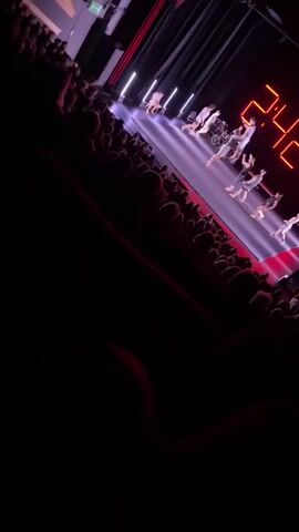 The video pans across a darkened theater audience, showing their backs to the camera, as they watch a performance on a brightly lit stage. On stage, a group of dancers are performing in front of a large red digital clock displaying the time 24:48:49. The stage is illuminated with purple and white lights, and there are black curtains in the background. The audience is seated in rows of red chairs. The video is shot from a high angle, looking down at the stage and the audience.