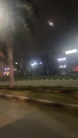 This is a blurry, nighttime video taken from inside a moving vehicle. The view is of a street scene with trees, streetlights, and buildings with illuminated signs on the right. The signs are difficult to read due to the blurriness and distance. The road is visible in the foreground with a curb and grassy median. There are a few vehicles visible on the road. The overall impression is of a commercial or urban area at night.