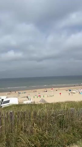 The video shows a view of a beach on a cloudy day. In the foreground, there is a wooden fence with tall grass growing behind it. A paved path runs alongside the beach, with people walking on it. The beach itself has sand, lounge chairs, umbrellas, and a few beachgoers. There are also some tents and structures on the beach, possibly beachside cafes or bars. The ocean is visible in the background, with waves gently lapping at the shore. The sky is filled with large, grey clouds, suggesting overcast weather. The overall impression is of a moderately busy beach on a day that is not particularly sunny.