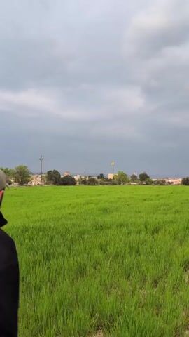 A man wearing a black sweatshirt, black pants, and a baseball cap runs through a lush green field towards a town in the distance. The sky is overcast. The man extends his arms, as if balancing, while running.