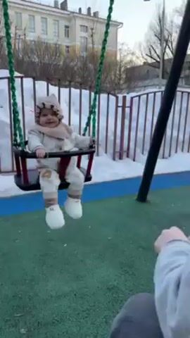 The video shows a young child, likely a baby, being pushed on a swing by a person who is only partially visible. The child is wearing a beige outfit including a hat and scarf, and is sitting in a red and black swing with green chains. The child is smiling. The person pushing the swing is wearing a grey sweater. The swing is located in a playground with a green and blue ground covering. There is snow visible in the background near a white fence and behind a building. The weather appears to be sunny.