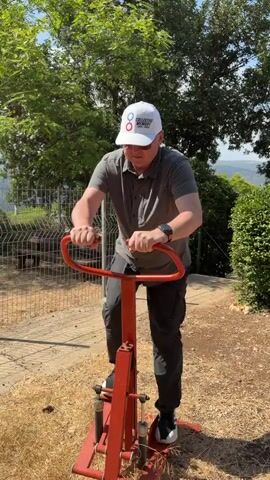 The video shows a man in his 40s or 50s exercising on an outdoor fitness machine. He is wearing a gray polo shirt, dark pants, white sneakers, and a white baseball cap with a logo that appears to say "COLLECTIVE MEMORY" and "THREE RIVERS". The machine is bright orange and appears to be a stepper or climber. The man is gripping the handlebars and appears to be stepping up and down.

The setting is outdoors, with dry, yellowish grass on the ground and a paved path visible in the background. There are trees and green bushes, suggesting a park or outdoor recreational area. A metal fence is also visible. In the distance, there are rolling hills and a bright sky, indicating it is daytime and likely sunny. The man's expression is neutral as he focuses on his exercise. The overall impression is one of an active person enjoying an outdoor workout in a natural, somewhat rustic environment. The time of day appears to be daytime, with clear lighting. The location context of Misgav Regional Council, Israel, suggests a Mediterranean climate, which is consistent with the dry grass and sunny appearance.