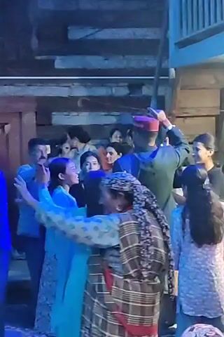 The video shows a group of people gathered outdoors at night. The lighting is dim and appears to be artificial, casting blue and white tones on the scene. Most of the individuals are facing toward the center of the group, suggesting they are engaged in a shared activity. Several people are wearing traditional clothing, including patterned shawls and caps. Some individuals are holding hands and appear to be dancing or participating in a communal gathering. One man in the center is wearing a dark jacket and a maroon cap, and he is facing away from the camera, with his arm raised as if pointing or gesturing. Other individuals in the background are partially obscured but seem to be observing or participating in the event. The environment appears to be rustic, with wooden beams visible in the background. The overall atmosphere suggests a celebratory or social event.