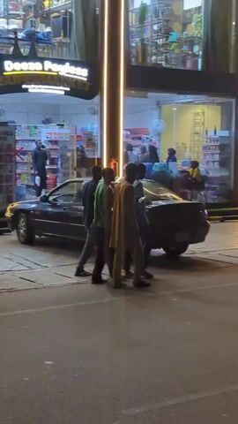 A group of four men walk along a city street. Two parked cars, a black sedan and a white SUV, are visible near them. The street is lined with shops, their windows brightly lit with fluorescent lights. The time is likely night, based on the darkness of the sky.