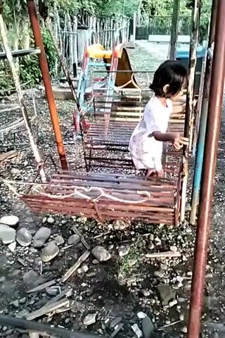 A young child is playing on a swing set in a backyard. The child, who appears to be a girl with dark hair, is wearing a light-colored t-shirt and shorts. She is standing on the seat of the swing, holding onto the metal frame as the swing moves back and forth. The swing set is old and rusty, with a wooden seat and metal frame. A heart-shaped design is visible on the front of the swing. Behind the swing set, there is a slide and some other playground equipment. The ground is covered in dirt, small rocks, and some scattered debris. The background shows trees and foliage, suggesting the area is residential or a park setting. The lighting indicates it is daytime.