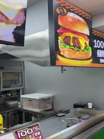 The video depicts the interior of a burger restaurant. The scene includes a display screen showing a burger, stainless steel refrigerators and counters, food preparation areas with sauce bottles, a tissue box hanging above a green point-of-sale terminal, and stacked cardboard boxes. The environment appears to be a fast-food kitchen or service area.