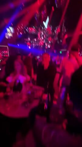The video shows a crowded nightclub interior with people milling around tables. Red and blue spotlights illuminate the scene. Disco balls hang from the ceiling, reflecting the light. A table is visible in the foreground with drinks and other items on it. Some people are looking at their phones.  Two men walk towards the camera near the end of the clip.