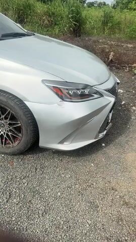 The video shows the front corner of a silver Lexus sedan parked on a gravel surface. The car has a dent on the front bumper and a scratch on the headlight. The wheel is visible and appears to be a custom silver alloy with red accents. In the background, there is green vegetation and a cloudy sky. The camera is positioned low and close to the car, focusing on the damaged front end. The footage appears to be taken during the daytime.