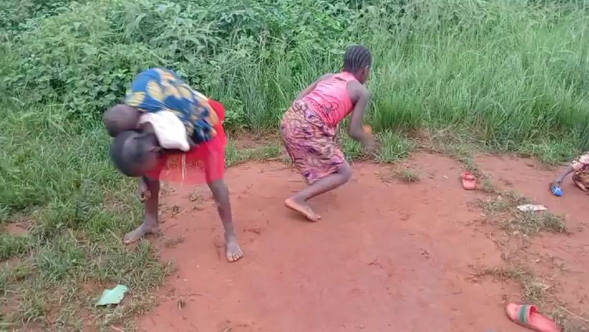 The video shows a group of children playing in a grassy area with a red dirt patch. One child carries a baby on her back in a fabric sling. Another child stumbles, and the carrier turns to look at her. The children are all dressed in casual clothing. Some of the children are sitting on the ground playing with small objects. The background is a blur of green vegetation.