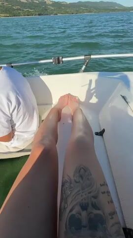 The video begins with a close-up of a person's legs and feet as they rest on the edge of a white boat. The person is wearing white shorts and has tattoos on their legs. The boat is on a body of water, likely a lake, with green trees and hills visible in the background. The sky is a clear blue. As the video progresses, the camera pans out to show more of the boat and the surrounding scenery. The water is calm and reflects the blue sky. The boat appears to be moving slowly, indicated by the slight ripples on the water's surface. Another person, wearing a white t-shirt, is partially visible in the left of the frame. The video ends with a wider shot of the boat moving across the water, with the shoreline and distant hills in view.