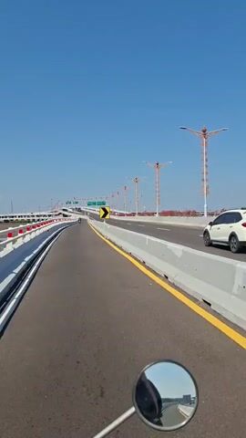 The video shows a first-person view from a motorcycle traveling on a highway under a clear blue sky. The road is paved and has a yellow line on the right edge. On the right side of the road, there is a concrete barrier and a white car is visible. On the left side of the road, there is a white railing. There are streetlights with decorative orange tops lining the highway. A yellow chevron sign indicating a curve to the right is visible. The motorcycle's circular rearview mirror is in the foreground, reflecting the road and sky.