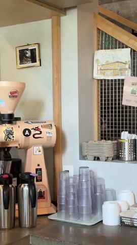 The camera pans up from a countertop loaded with plastic cups, paper cups, and a silver thermos to reveal a coffee grinder adorned with stickers. Next to it, there is an espresso machine and some shelves stocked with various coffee-related items. Above, wine glasses hang upside down from a rack. A framed photograph and a print that reads "PROUDLY SERVING ANCHORED COFFEE" hang on the wall. In the final frame, a woman's hand appears as she approaches the counter.